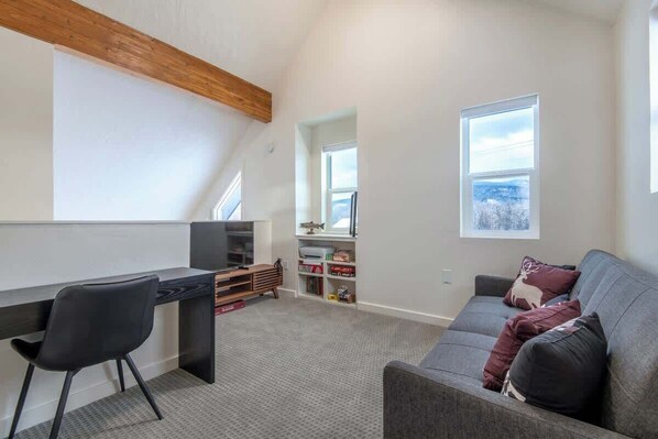 A modern loft bedroom with a desk, a grey sofa, and a large window offering a view of the outdoors. - Teton Homestead: Rocking Chair Ski Retreat (Driggs)