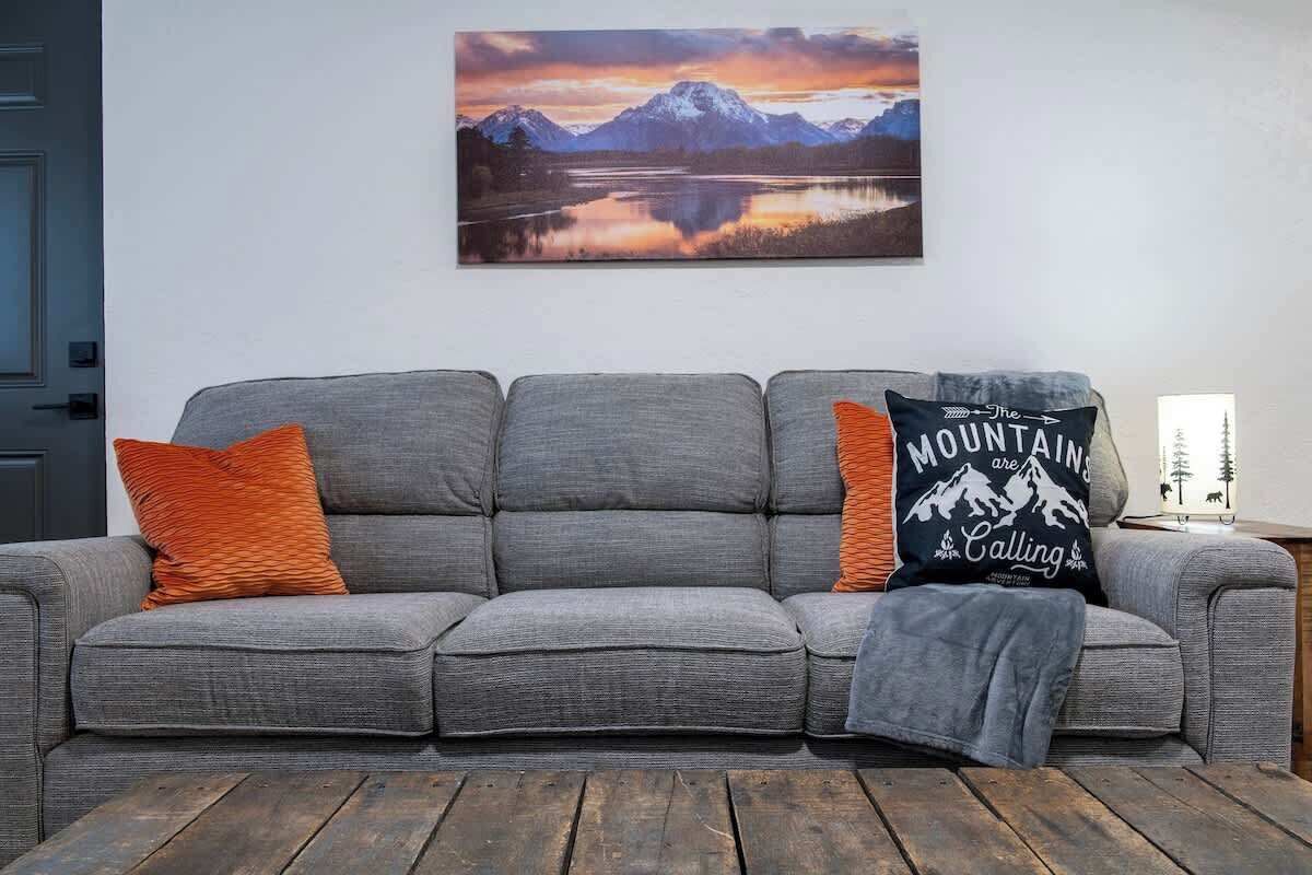 Un sofá gris con dos cojines naranja y uno negro, una mesa de centro de madera y un cuadro de un paisaje montañoso en la pared.