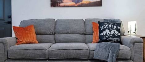Un sofá gris con dos cojines naranja y uno negro, una mesa de centro de madera y un cuadro de un paisaje montañoso en la pared.