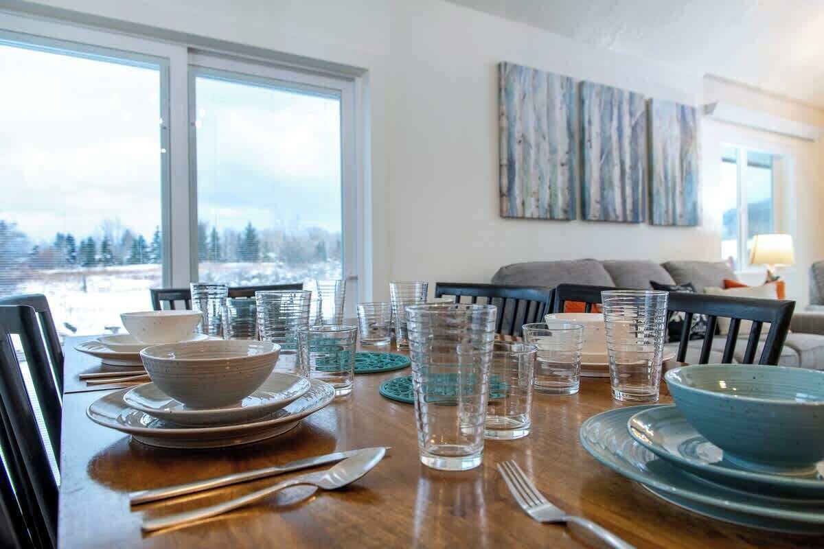 A dining table set with plates, bowls, and glasses, with a view of trees through large windows.