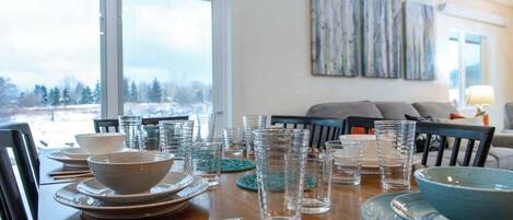A dining table set with plates, bowls, and glasses, with a view of trees through large windows.