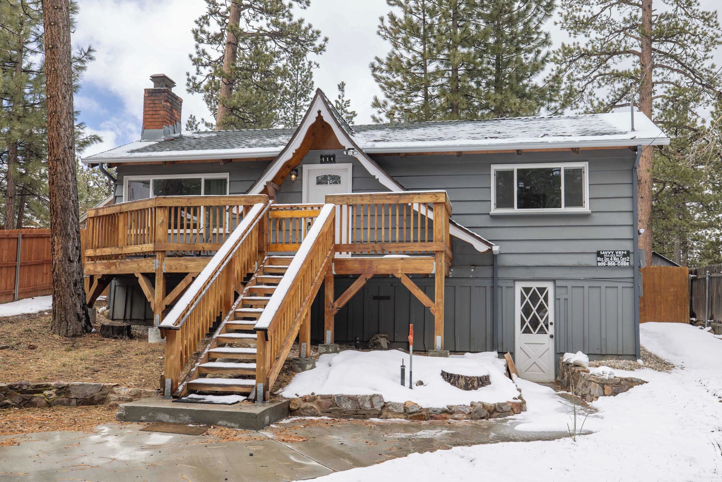 A two-story cabin with a wooden deck and a stone foundation, surrounded by trees.