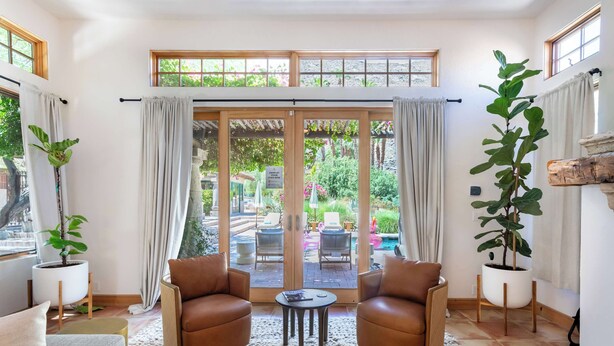 Un salon moderne avec deux fauteuils en cuir, une petite table ronde et une grande porte-fenêtre coulissante donnant sur une terrasse extérieure équipée de chaises et d’une table.