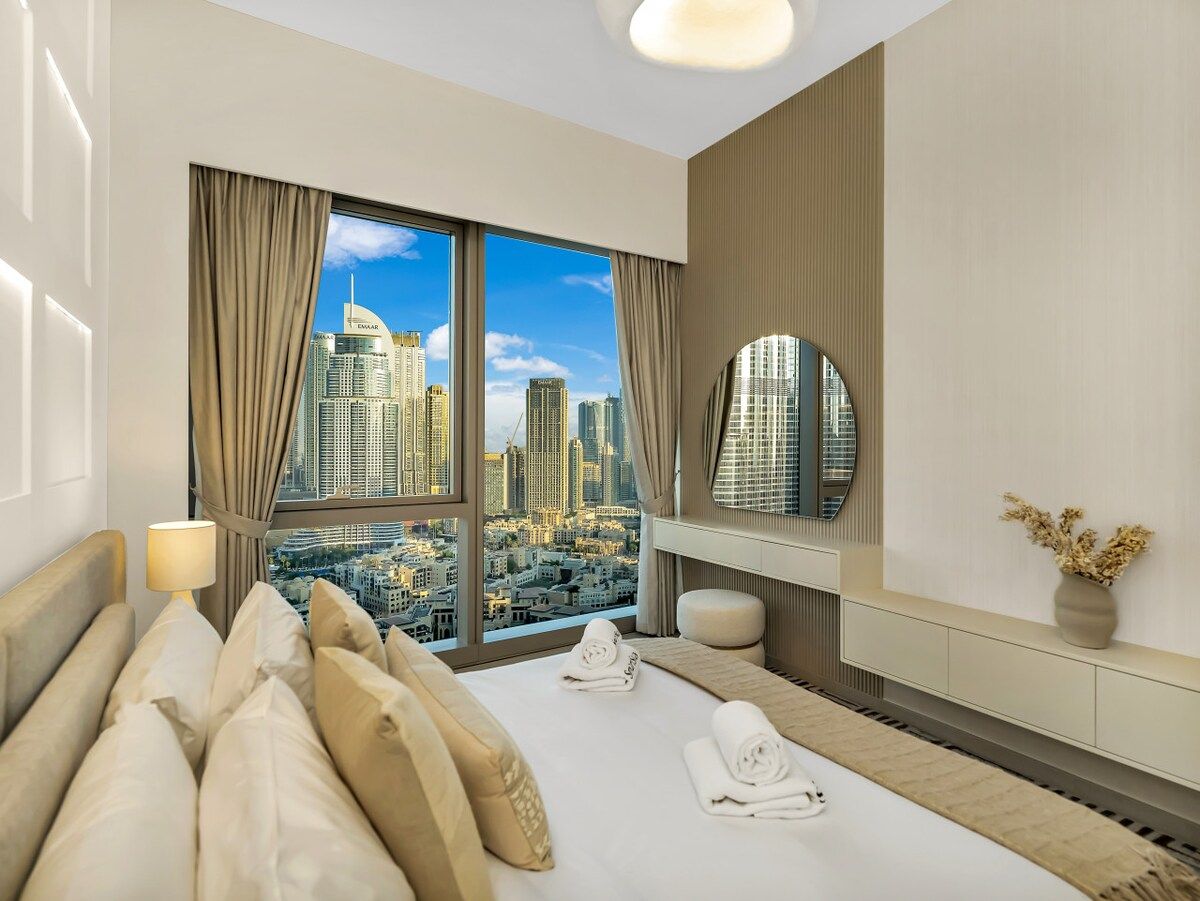 A hotel room with a large bed, a bedside table with a lamp, a mirror, and a view of a cityscape.