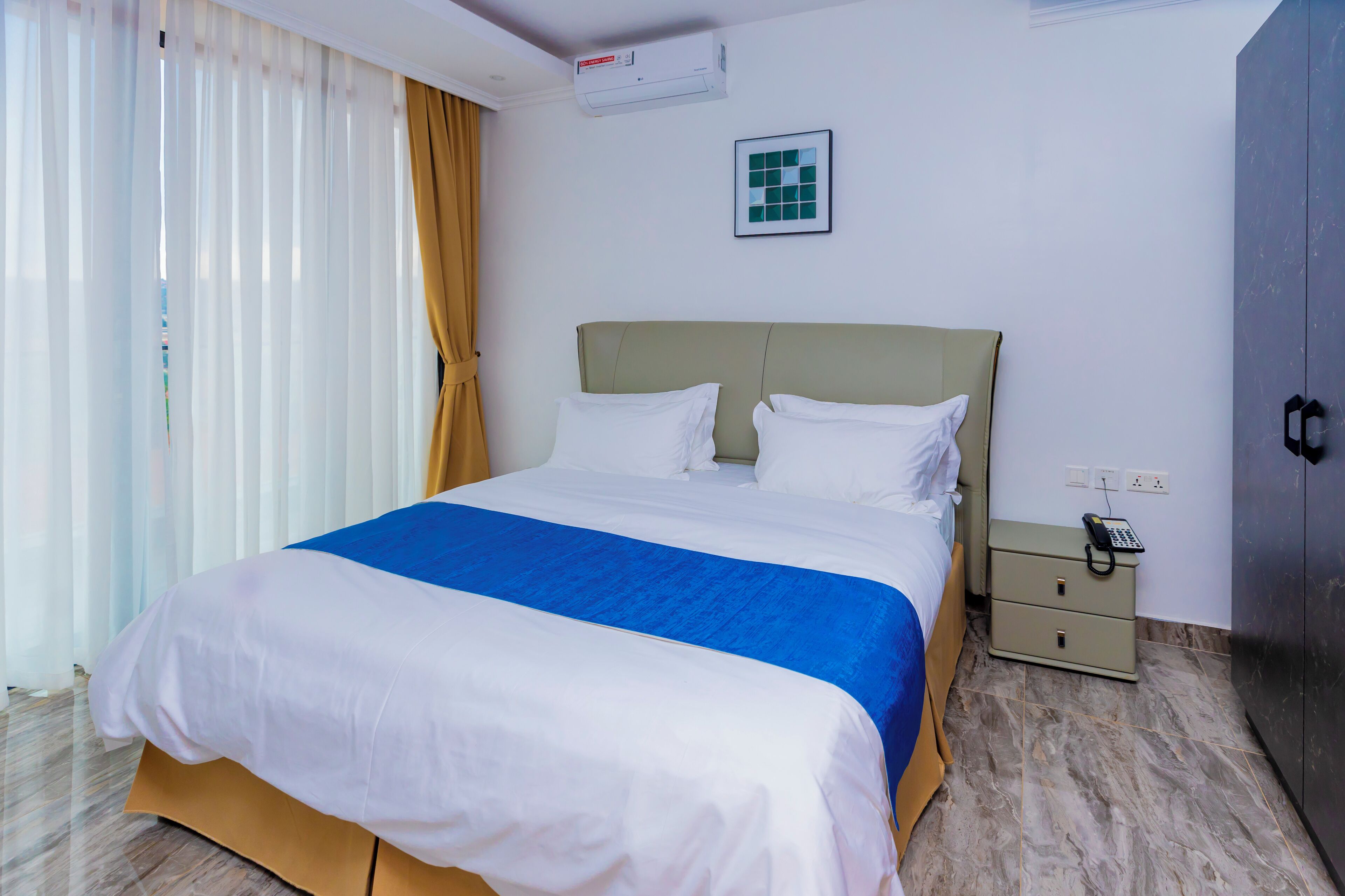 A hotel room with a bed, a nightstand, an air conditioning unit, a wall-mounted phone, and a wardrobe.