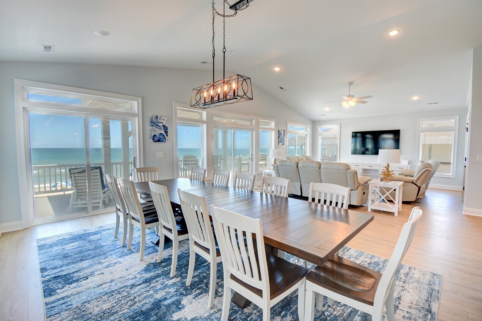 A spacious living area with a large wooden dining table surrounded by white chairs, a sofa, and a TV.
