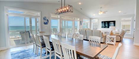 A spacious living area with a large wooden dining table surrounded by white chairs, a sofa, and a TV.