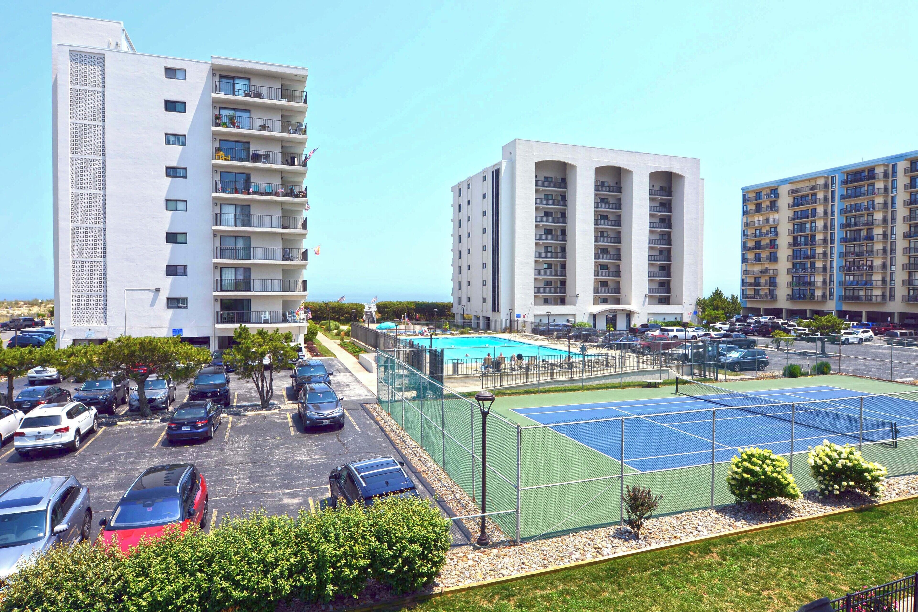 Ein mehrgeschossiges Parkhaus mit mehreren Autos, einem Tennisplatz und einem Poolbereich, umgeben von Gebäuden.