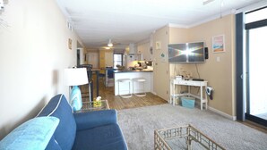 Una sala de estar con un sofá azul, un televisor montado en la pared y una cocina al fondo.
