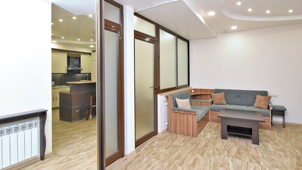Un salon moderne avec un canapé d’angle, une table basse et un coin cuisine visible à travers une porte vitrée.