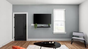 A modern living room with a flat-screen TV mounted on the wall, a wooden floor, a black coffee table with decorative items, a grey armchair, and a white door.