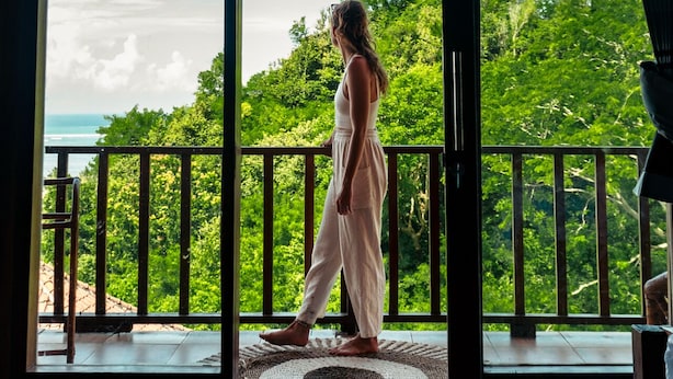 A woman walking through a glass door onto a balcony with a view of a forest and ocean.