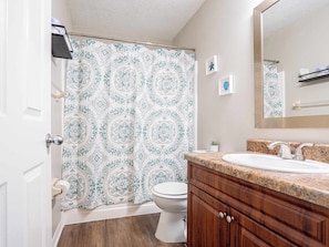 Un baño con cortina de ducha estampada, un inodoro, un lavabo con encimera de granito, armarios de madera y un espejo.