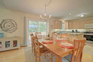 A dining area with a wooden table set for four, surrounded by chairs, a chandelier, and a view through a large window. - Spacious home with deck and fire pit! (Brewster)