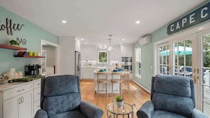 Une cuisine moderne avec des meubles blancs, un îlot central et un coin repas comprenant deux fauteuils bleus et une table basse en verre.