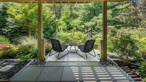 En træpergola med to sorte fletstole og et lille bord, omgivet af en have med forskellige planter og en stensti.