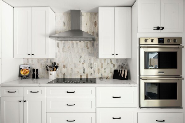A kitchen with white cabinets, a stainless steel range hood, a double oven, and a backsplash with a geometric tile pattern.
