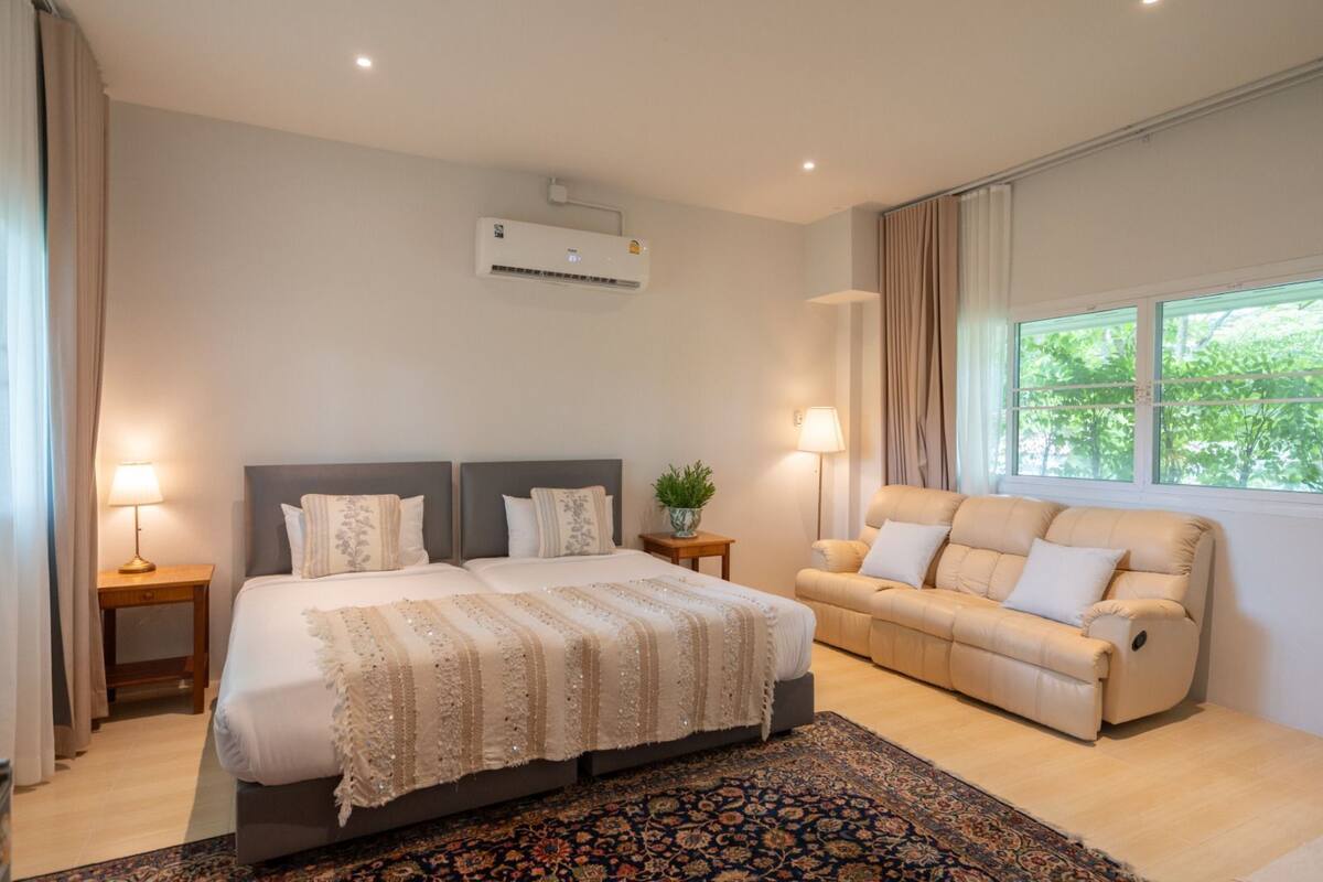 A bedroom with a large bed, a sofa, a small table with a lamp, and a window with curtains.