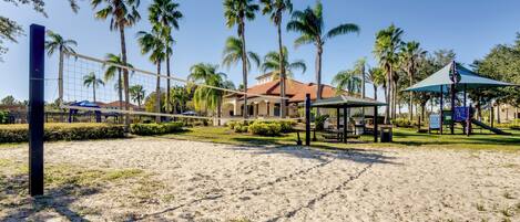 A beach volleyball court with palm trees, a gazebo, and a playground in the background.