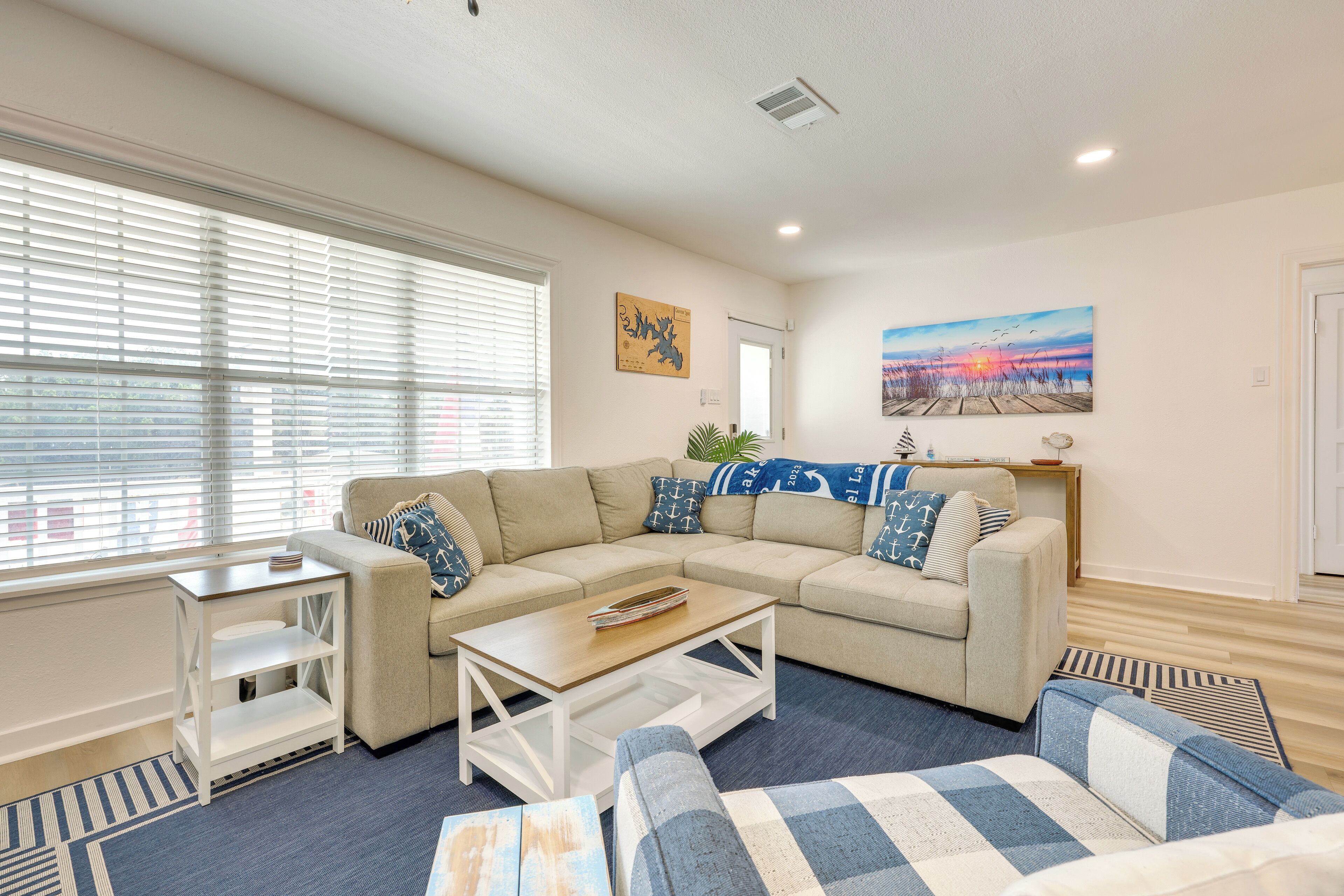 A living room with a beige sectional sofa, a coffee table, and a blue and white patterned rug. There are two side tables, one with a lamp and the other with a plant.
