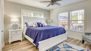 Un dormitorio con cama, mesitas de noche, ventilador de techo y ventanas amplias.