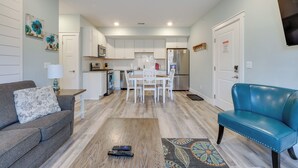 Un salon moderne avec un canapé gris, un fauteuil en cuir bleu et une table basse en bois. On aperçoit à l’arrière-plan une cuisine avec des meubles blancs et un coin repas comprenant une table et des chaises blanches.
