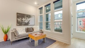 A modern living room with a grey sofa, a wooden coffee table, a large abstract painting, and large windows offering a view of buildings and trees. - Near Downtown: Luxury Townhome in Bentonville! (Bentonville)