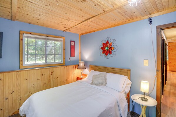 Een slaapkamer met een houten plafond, een raam met uitzicht op bomen, een bed met wit beddengoed, een nachtkastje met een lamp en een wanddecoratie.