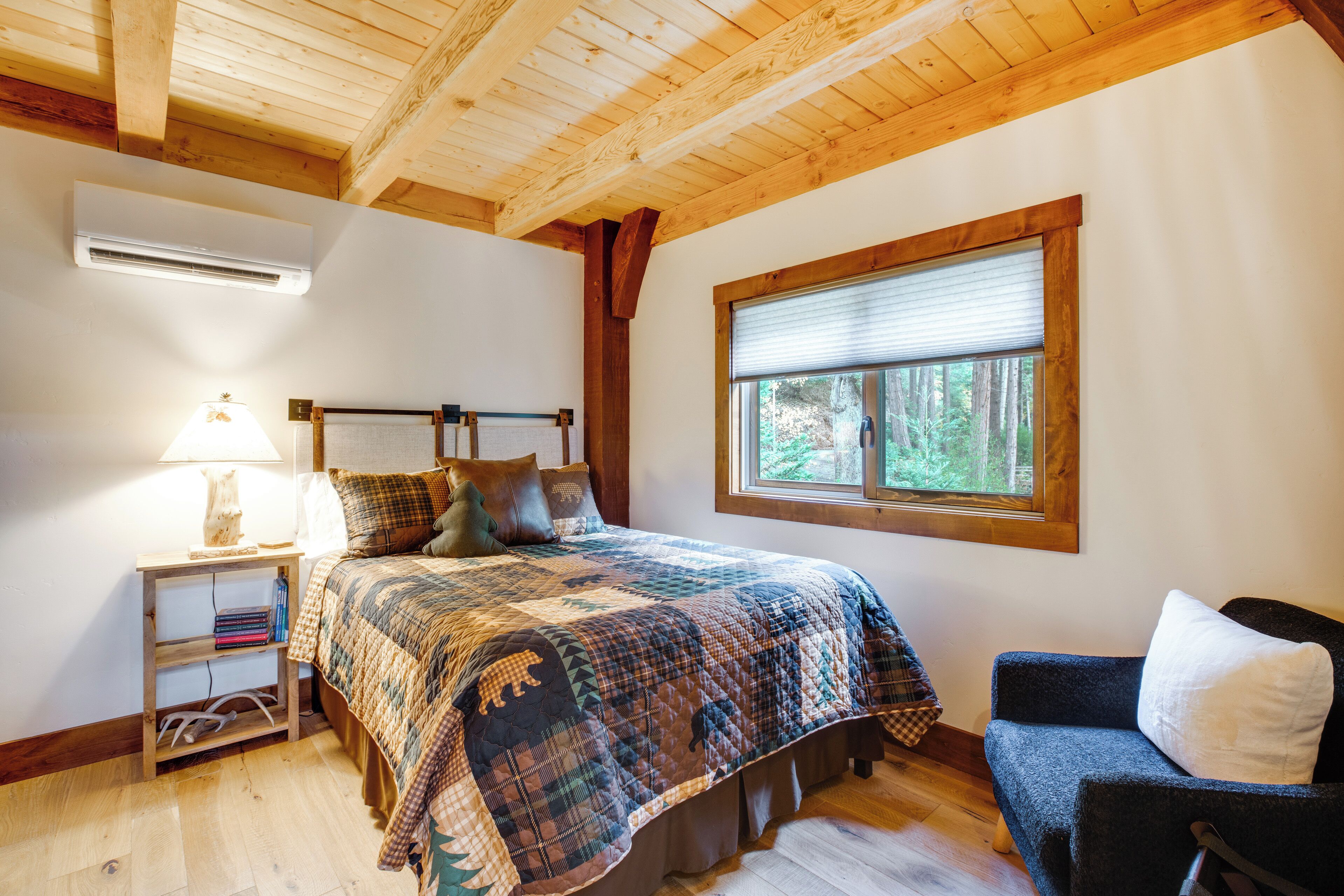A bedroom with a wooden ceiling, a bed with a quilt, a nightstand with a lamp, a window with blinds, and a chair.