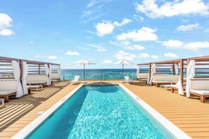 Un área de piscina con agua azul cristalina, terraza de madera, sillas de descanso blancas y sombrillas.