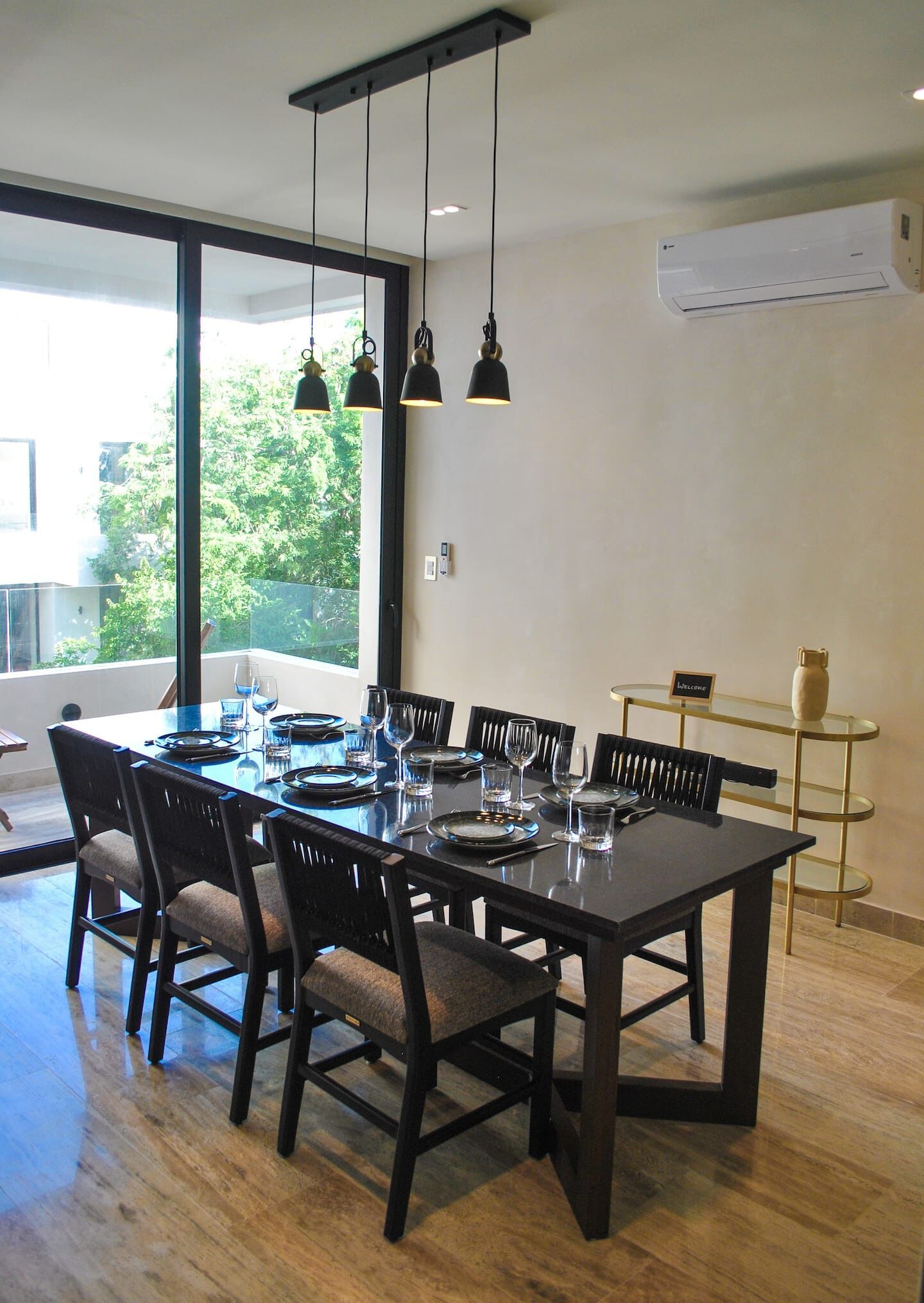 Un espace repas moderne avec une grande table rectangulaire dressée pour six personnes, entourée de chaises en bois foncé. Trois lampes suspendues sont disposées au-dessus de la table.