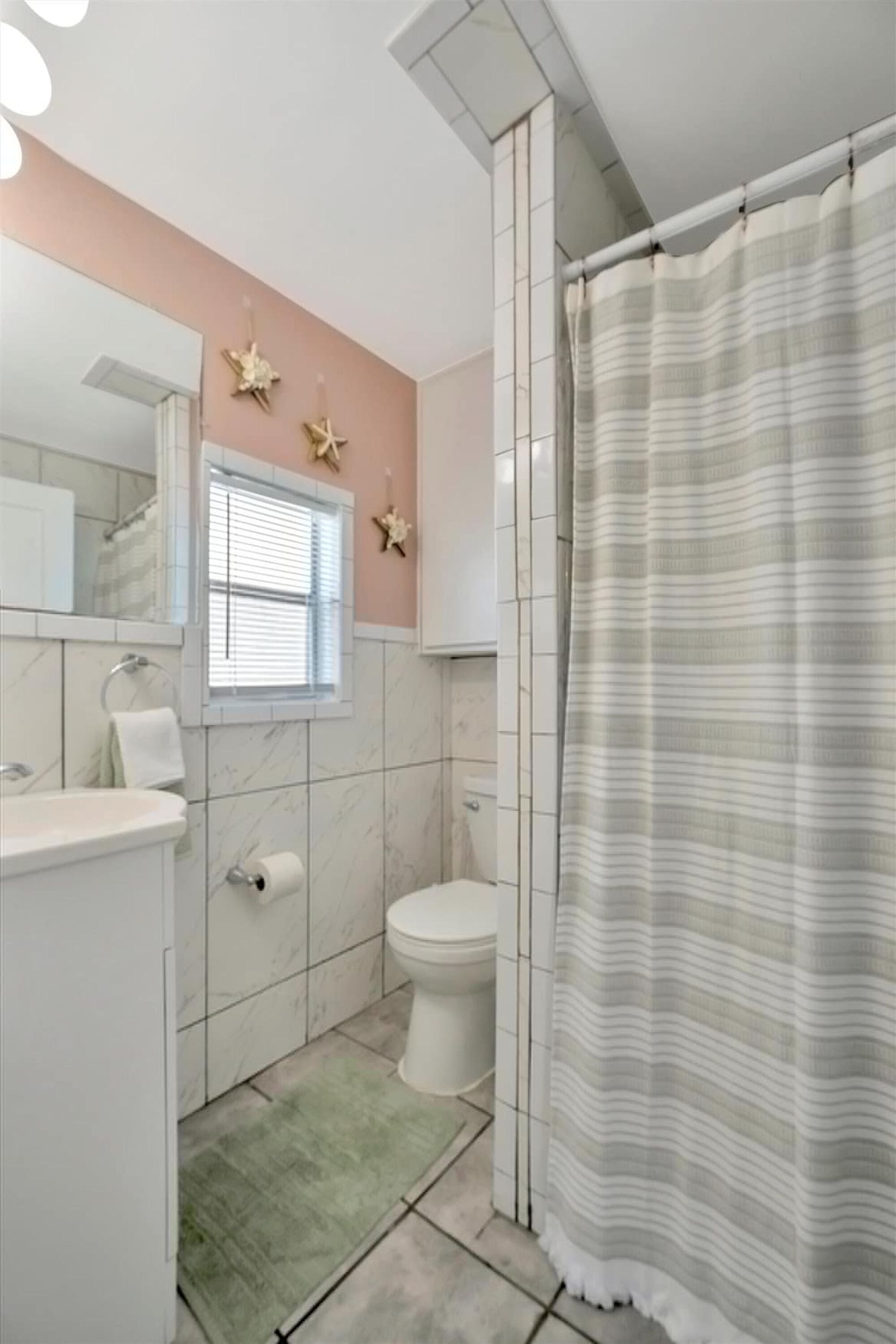 A bathroom with a toilet, sink, and mirror. There is a window with blinds, a green rug, and a shower curtain.