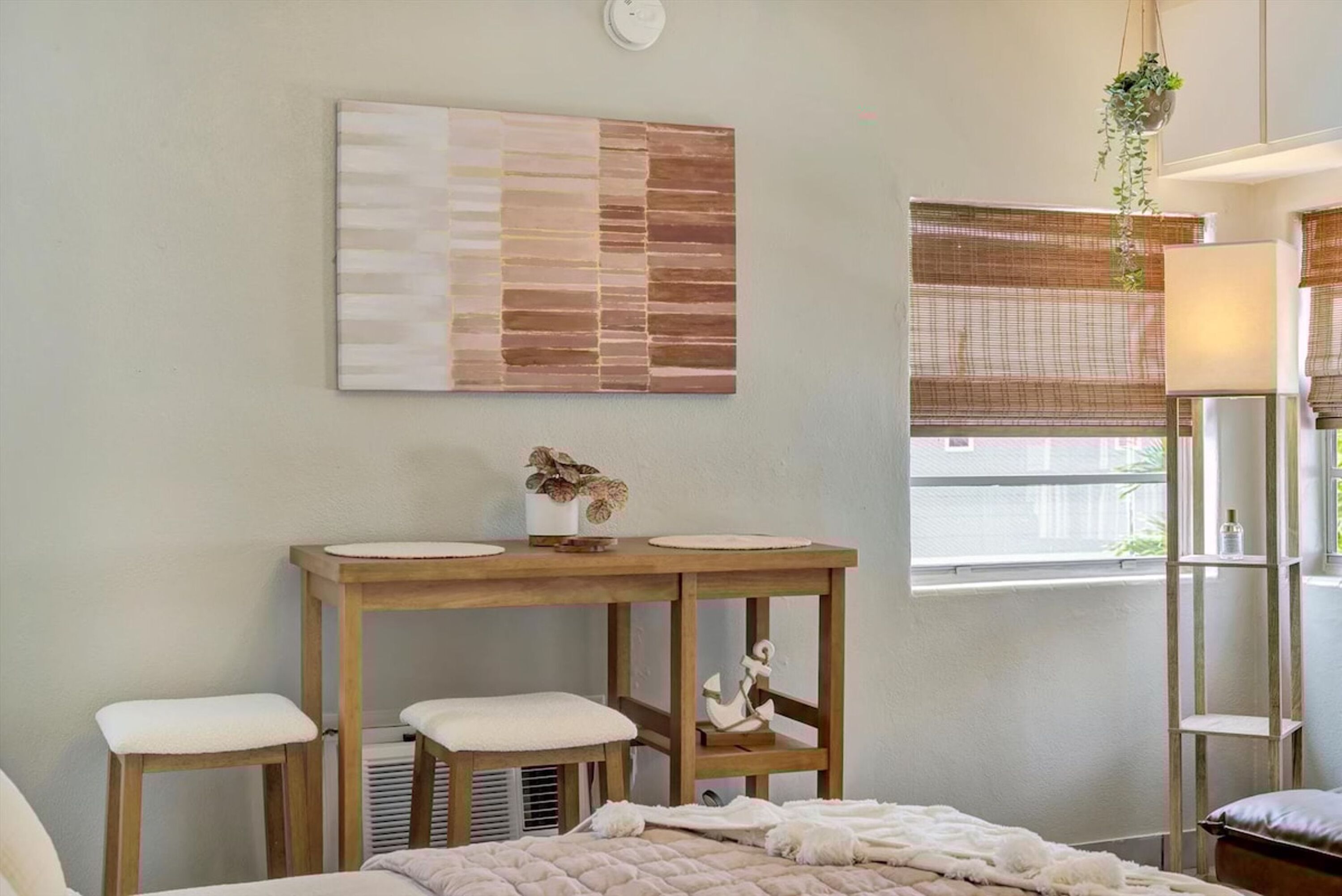 A bedroom with a bed, a wooden table with stools, a wall-mounted artwork, and a window with blinds.