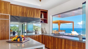 A modern kitchen with wooden cabinets, a large island with fruit, and a view of the sea through a glass door. - Villa Amandara – Iconic Luxury Estate in St. Martin (Les Terres Basses)