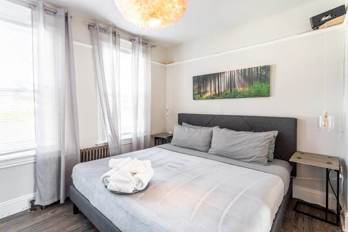 A modern hotel room with a large bed, a headboard, a bedside table, a nightstand, a wall-mounted artwork, and a window with curtains.