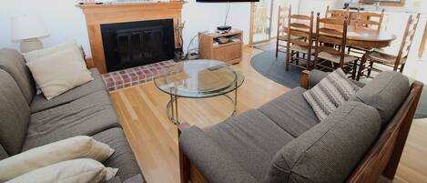 A living room with a sofa, a glass coffee table, a fireplace, a TV, and a dining area with a wooden table and chairs.