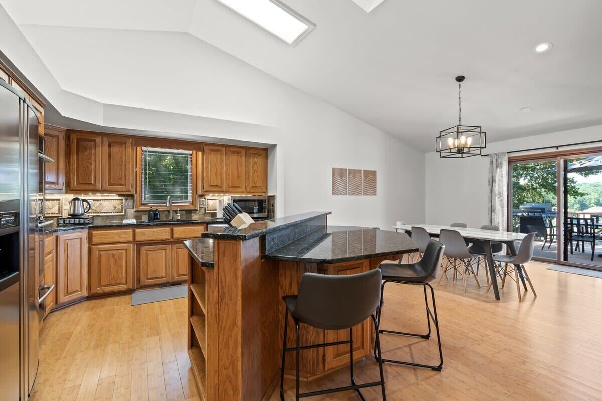 A modern kitchen with wooden cabinets, a central island with bar stools, and a dining area with a large table and chairs.