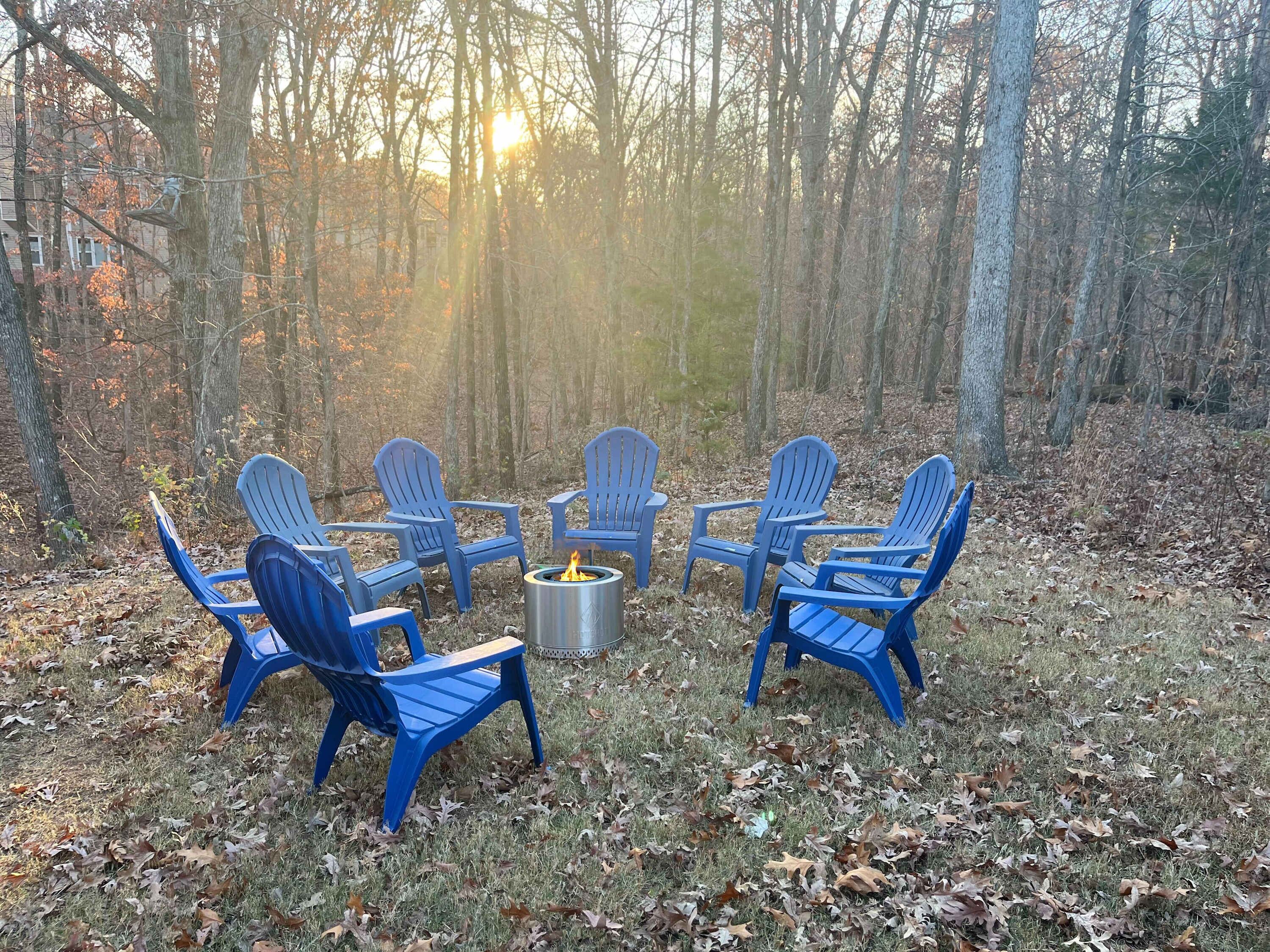 A circular arrangement of blue Adirondack chairs around a metal fire pit in a forested area.