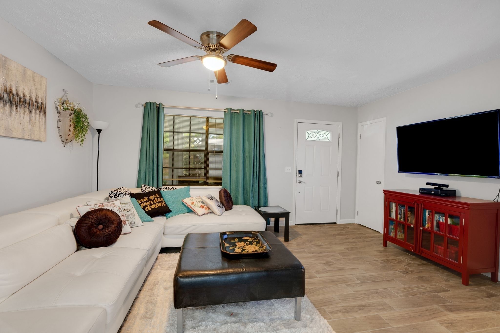 A living room with a white sofa, a black ottoman, a ceiling fan, a television, and a red cabinet.