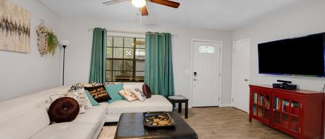 A living room with a white sofa, a black ottoman, a ceiling fan, a television, and a red cabinet.