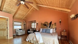 Un dormitorio con techo inclinado, suelo de madera, cama con ropa de cama estampada, mesita de noche, una silla, ventana con persianas y una lámpara de pared.