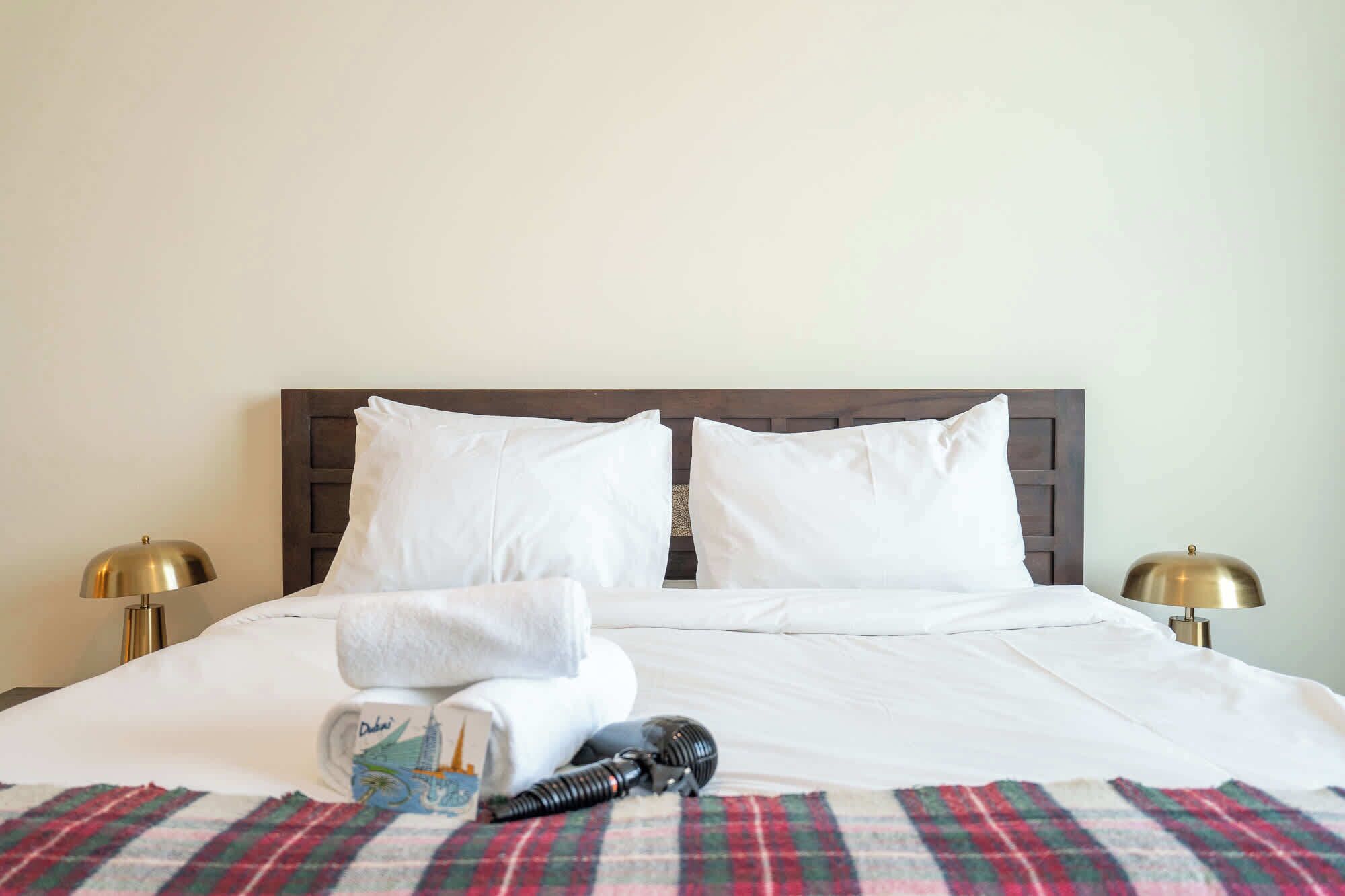 A neatly made bed with white linens, two pillows, a folded towel, a small card, and a hairdryer on a plaid blanket.