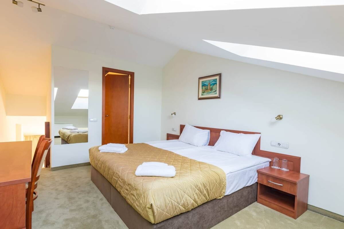 A hotel room with a large bed, a wooden nightstand, a desk, and a mirror.