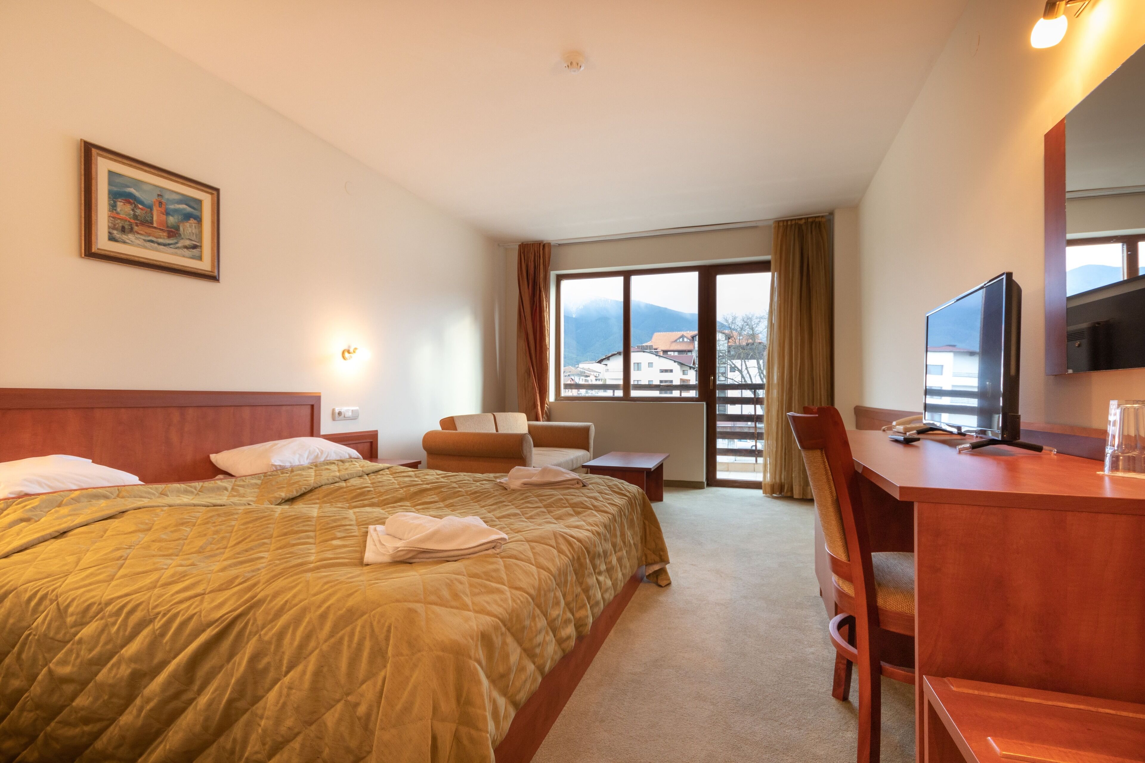 Une chambre d’hôtel équipée d’un lit, d’un bureau, d’une chaise, d’une télévision et d’une fenêtre donnant sur des bâtiments et des montagnes.