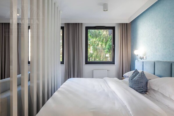 A hotel room with a large bed, a window with a view of greenery, a radiator, and a light fixture.