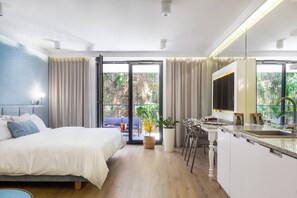 A modern hotel room with a bed, a dining area, a kitchen, and a balcony with a view of greenery.