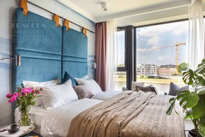 Une chambre d’hôtel moderne avec un grand lit, une tête de lit dotée d’un panneau rembourré bleu, une vue sur un chantier de construction et une plante en pot.