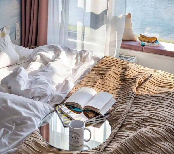 A bed with white linens, a glass table with a book and mug, and a window with curtains.