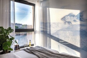 Hotellihuone, jossa on suuri ikkuna, sänky, huonekasvi sekä näkymä joelle ja rakennuksille.