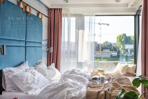 A bedroom with a large bed, a headboard, a window with curtains, and a view of a construction site.